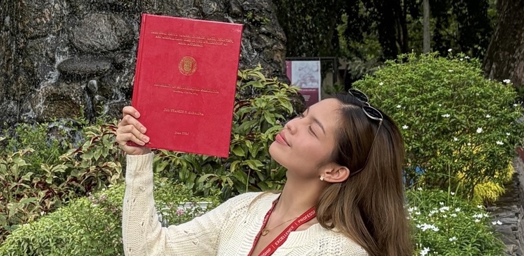Jill Frances Cabaluna holding her hardbound undergraduate thesis in front of the University of Southeastern Philippines seal